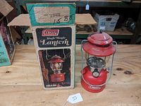 Full view of red Coleman single-mantle lantern next to its original box on wooden surface.