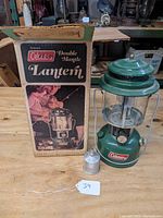 Green Coleman lantern with original box and fuel canister on wooden surface, showing branding and model number on the box and lantern base.