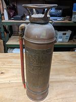 Full view of copper fire extinguisher standing upright on wooden surface with items in background.
