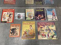 Full lot photo showing all 8 books and a framed print laid out on a metal floor.
