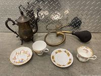 Photo showing two decorative teacups, three decorative plates including one with Asian motifs, an ornate metal teapot, and a vintage brass bulb horn with black rubber bulb.