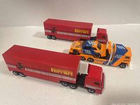 Two red Ferrari semi truck trailers with cab and one orange and blue Matchbox Hardee's semi truck carrying race car, arranged on white surface