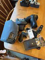 Photo showing various camera accessories arranged on wooden table including camera cases, Vivitar flash, binoculars, and other items.