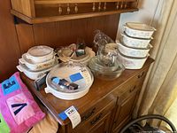 Overall view of 17 pieces of Corningware and glassware arranged on wooden furniture.