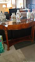 Full view of wood foyer table with curved scalloped top edge, decorated carved legs and lower shelf, shown with decorative vases on top.