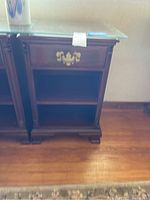 Front angle of wooden nightstand showing glass top, single drawer with brass handle, and lower shelves