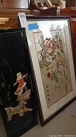 Shows three framed silk wall art panels leaning against a piano, one large colorful piece and two narrow black panels with stitched figures and calligraphy.