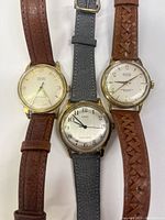 Photo showing Gruen Precision mechanical wristwatch with white face, gold-tone case, and brown leather strap alongside two other watches (Timex and Rutina).
