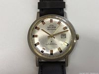 Front view of watch showing silver dial, gold-tone markers and hands, date window, and black leather strap.