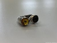 Close-up photo of a sterling silver ring featuring two round amber stones, yellow and dark brown, displayed on a neutral surface.