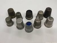 Front row and back row view of ten metal sewing thimbles on neutral surface showing varying textures, sizes, and designs including blue top insert on one thimble.