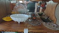 Rear view of milk glass bowl, clear glass egg plate, green serving dish, clear platters, small glass cups, and black metal basket with glass inserts displayed on shelf with wood backing and mirror.