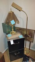 Photo showing black two drawer metal file cabinet with chrome handles, green metal file box, brass table lamp with amber glass panels on top, adjustable brass floor lamp to the right, and framed art print leaning against wall.