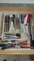 Top drawer with assorted flatware including forks, spoons, knives with plastic and stainless handles, plus small kitchen tools and a boxed Chicago Cutlery knife.