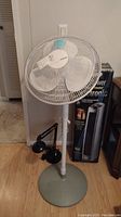 Holmes standing fan with white blades and protective grille, standing on a round metallic base in a room.