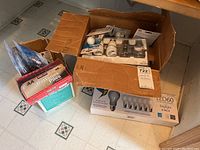 Two boxes containing light bulbs and batteries on tiled floor. One box open with various items including modern and older bulbs and batteries in packaging.