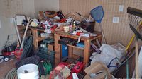 Photo of shed corner showing wooden workbench holding various yard tools, hardware, garden supplies, boxes, extension cords, and miscellaneous items.