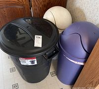 Three plastic lidded trash cans side by side: black large one, purple and cream smaller ones.