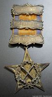 Front side showing star-shaped silver medal with laurel wreath and crossed tools, three engraved plaques bearing initials and numbers attached above, hanging from an orange and purple ribbon.