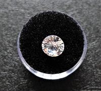 Top view of a clear cubic zirconia gemstone resting on black velvet inside a circular clear container, showing the facets and brilliance of the stone.