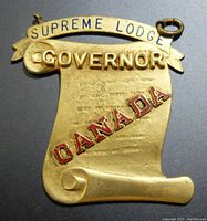 Front view of gold-tone medal shaped like a scroll with raised letters and red enamel "CANADA"