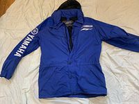 Blue Yamaha Factory Racing jacket with hood laid flat on a white sheet, showing front view with logos on chest and sleeve.