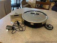 Photo showing Bobsweep robot vacuum cleaner, charging dock with lid open, power adapter, and remote control on a table.
