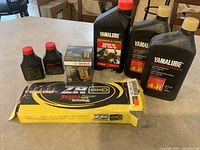 All items arranged on table: Regina motorcycle chain in box, Bosch oil filter box, three large Yamalube oil bottles, two small Sears Craftsman oil bottles.
