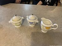 Photo showing three Noritake china pieces: one yellow creamer pitcher, one blue sugar bowl with lid, one yellow sugar bowl with lid on a countertop.