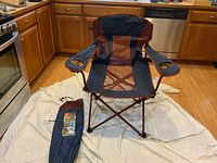 Blue and maroon oversized mesh Ozark Trail camp chair with two cup holders, set up on a white sheet in a kitchen. Accompanied by matching carry bag placed beside the chair.