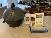 Photo showing gray portable charcoal grill with dome lid and air vent, two full bottles of lighter fluid, and a plastic container with assorted firestarter packs on a countertop.