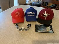 Photo shows three baseball caps, racquetball goggles, and set of darts on a table.