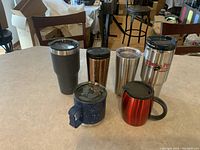 Photo of six travel beverage mugs on a countertop showing a variety of brands and colors including blue speckled, red Starbucks, pinkish Starbucks, two Ozark Trail, and a Stanley mug with logo.