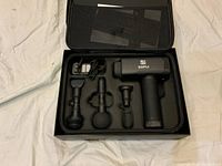 Opened box showing Supli black massage gun, four attachments, charging cable arranged neatly in molded black foam insert inside carry case.