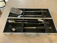 Four BBQ tools inside open metal carrying case arranged in custom slots on a beige countertop.