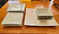 Top view of square white dinner plates, side plates, bowls, and large charger plate arranged on wood surface.