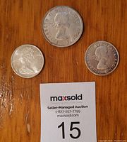Three Canadian coins on wood background with MaxSold auction card. Coins show Queen Elizabeth II on obverse.