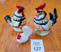 Two vintage ceramic rooster pitchers with removable lids and a white ceramic hen salt shaker on a wood surface with auction tag
