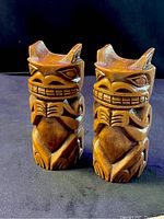 Two small wood carvings side by side on black surface, showing front and angled views of similar figures with expressive facial features.