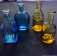 Four mini vintage Wheaton glass bottles, two blue and two amber, showing their distinct shapes and embossed details on a dark surface.