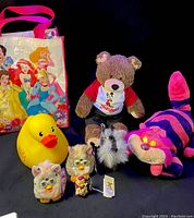 Photo showing assortment of stuffed animals and toys including Furby toys, Disneyland bear, rubber duck, Cheshire Cat plush, and princess tote bag.