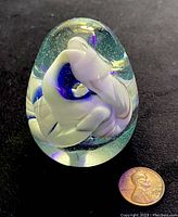 Full view of the egg-shaped glass paperweight with internal white and blue swirl design shown next to a penny for size reference.