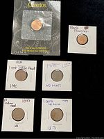 Five US pennies laid out showing dates and types plus a blank planchet in individual white paper holders.