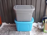 Two storage bins stacked, gray bin on top of a blue bin, both with lids, placed on a mat with some snow outside.
