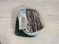 Photo showing mixed drill bits inside a plastic storage container, including some new ones still in packaging and several used bits.