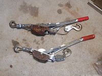 Two come along cable puller winches laid flat side by side showing top side with red handles, metal body, and hooks.