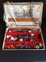 Top view of an open jewelry box showing red velvet compartments filled with assorted costume jewelry and a white satin lid with decoration. Visible a white beaded necklace, a metal choker, various earrings, pins, necklaces, and a small wristwatch.