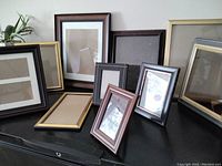 Various picture frames propped against a dark surface with plants in the background, showing different sizes and colors.