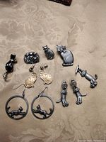 Collection of cat themed pins and earrings on a beige background cloth. Includes black cat pins with jewel accents, pewter hanging cat earrings, gold tone cat earrings, and pins with various cat poses including sitting and waving cats.