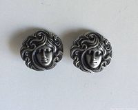 Two pewter buttons on a white background showing detailed female faces with flowing hair in Art Nouveau style.